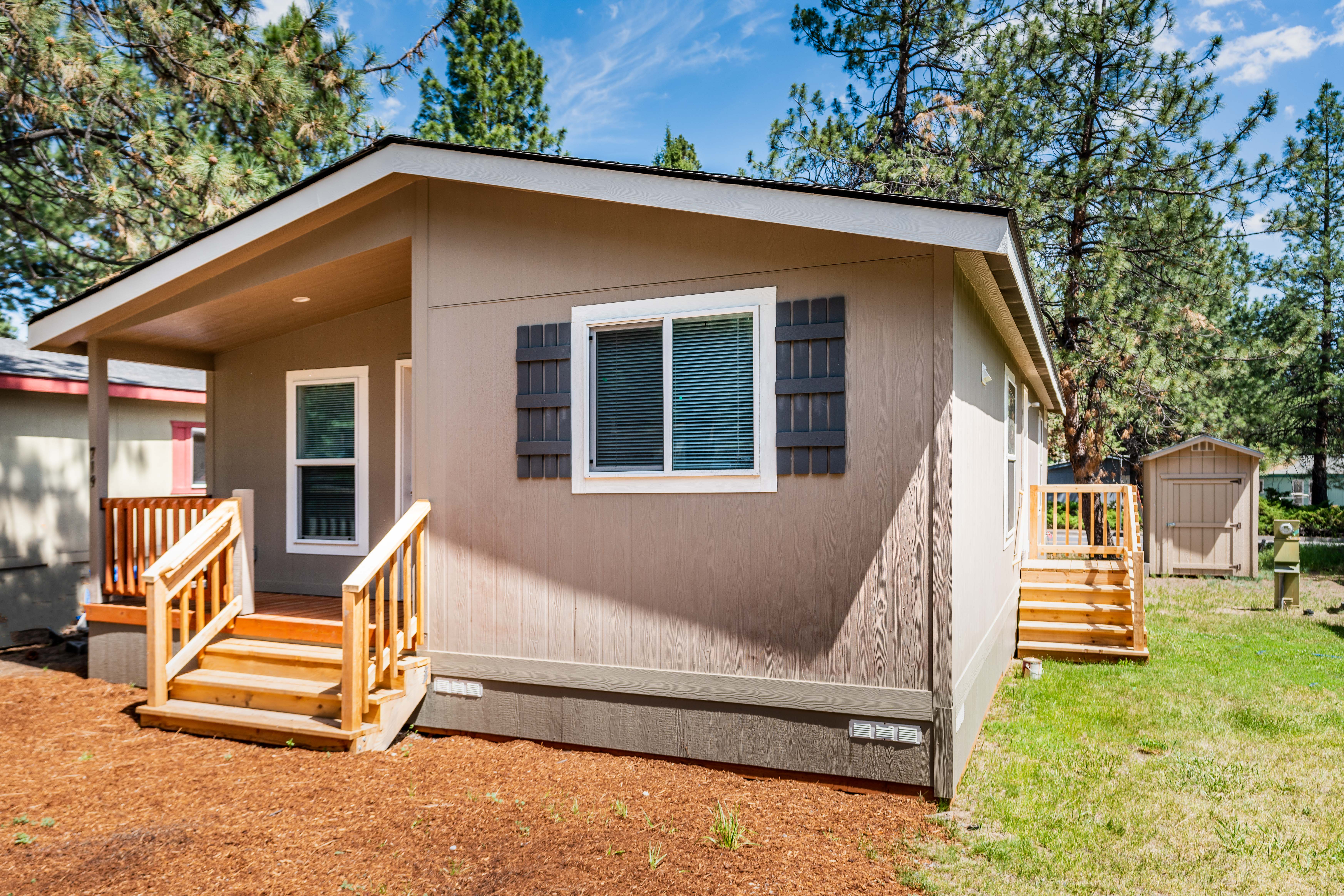 A small beige manufactured home with front steps, a window with shutters, and a storage shed in the backyard, surrounded by trees and grass.