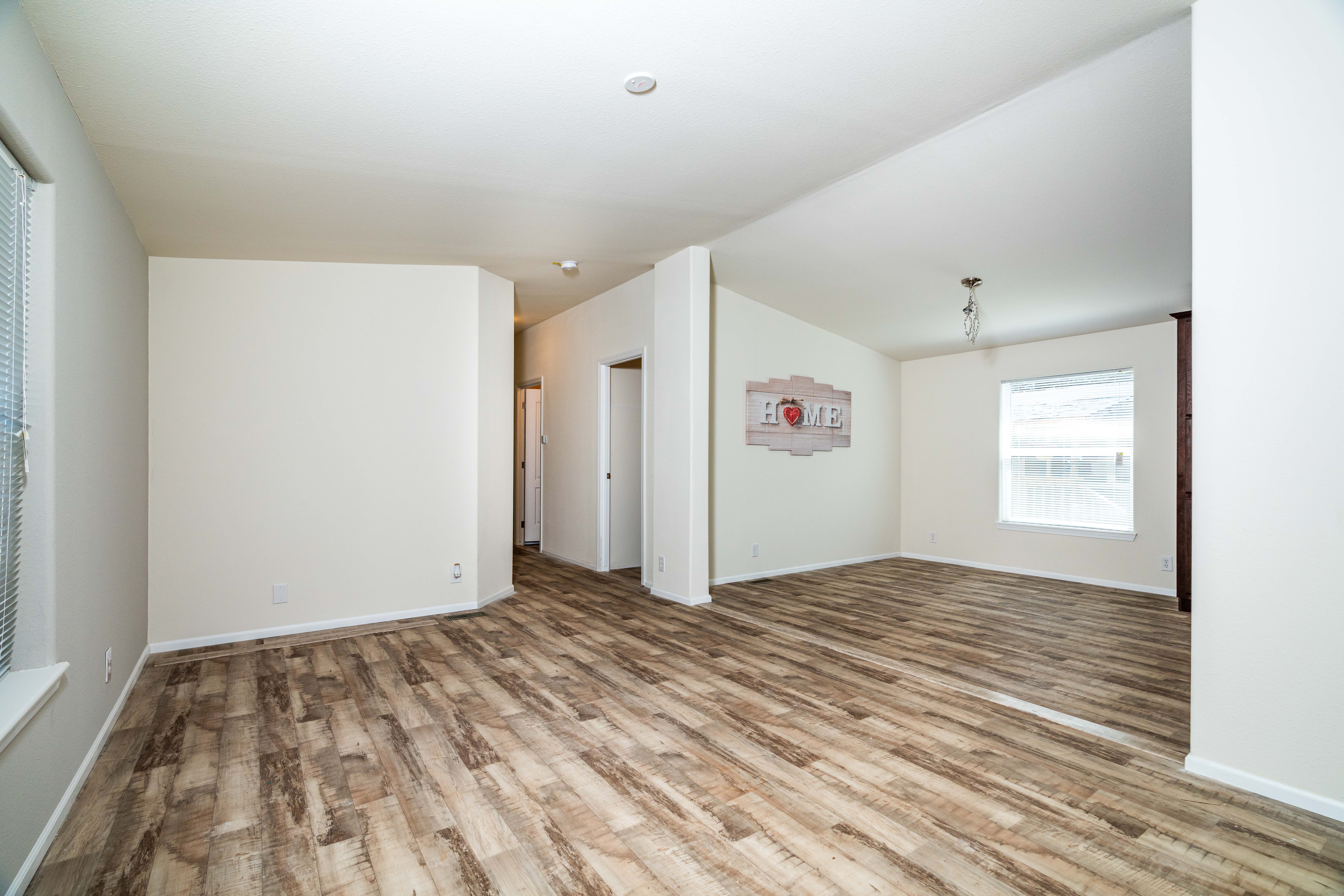 Empty living room with wood-look flooring, white walls, two windows, and a wall art piece reading "LOVE"; hallway leads to other rooms.