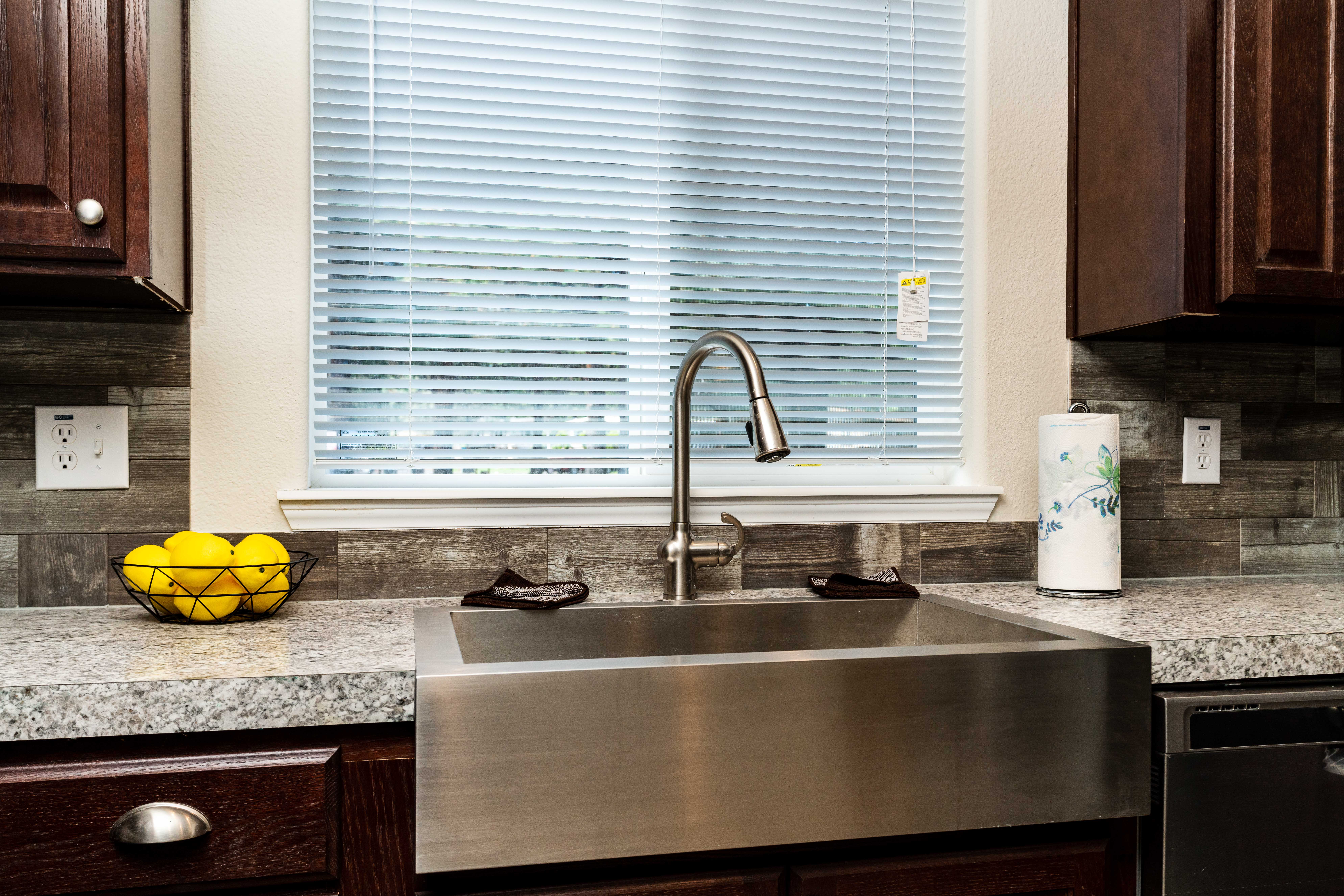 A modern kitchen sink with a stainless steel basin, a gooseneck faucet, a bowl of lemons, a paper towel roll, and a window with blinds above the counter.