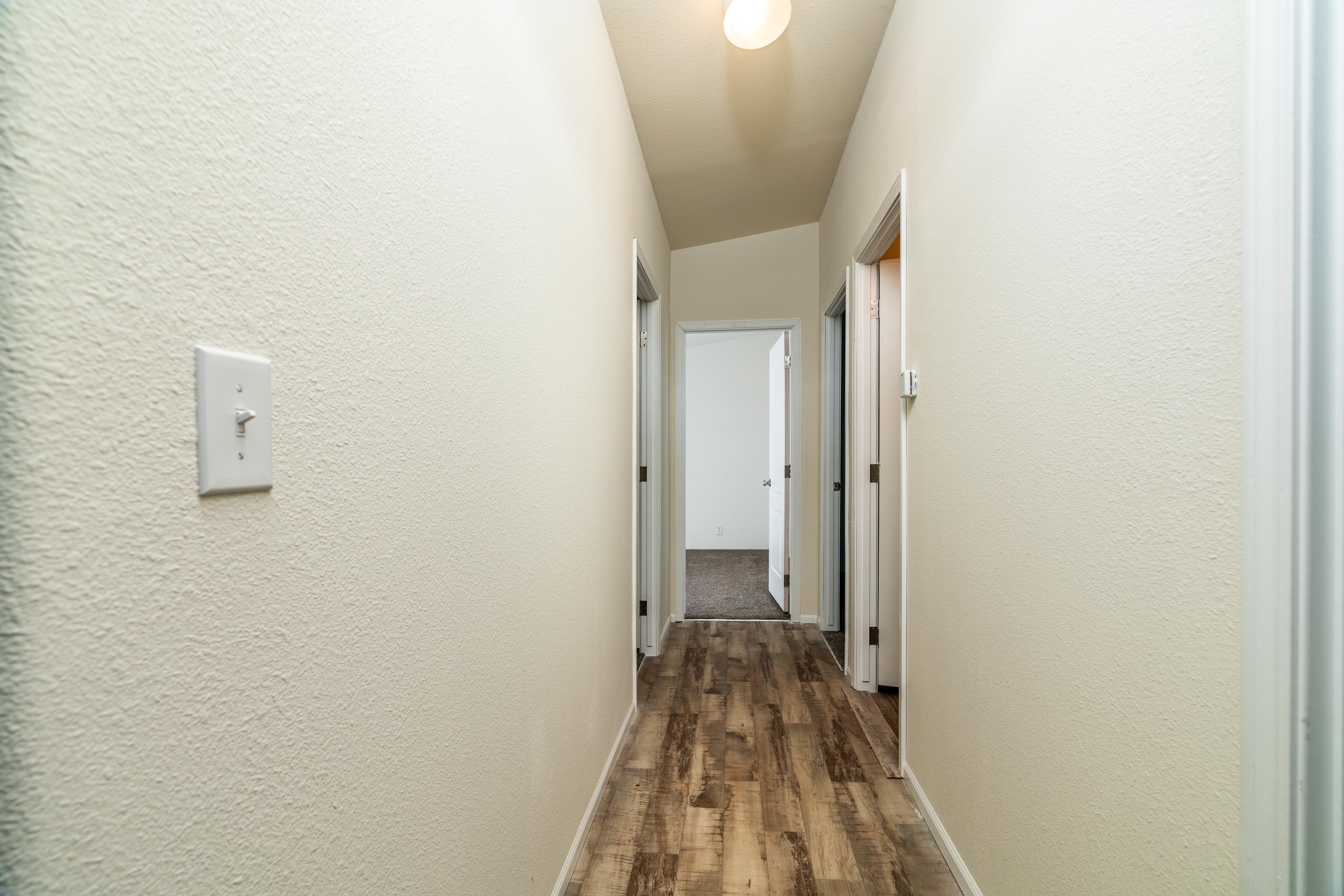 A hallway with light-colored textured walls, wood-style flooring, and four doorways leading to separate rooms, illuminated by a ceiling light.